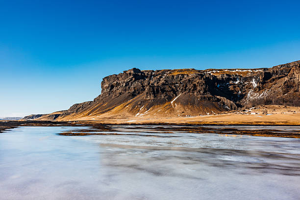 Expansive Icelandic landscape featuring a large rugged mountain plateau, golden grass fields, and a partially frozen lake with ice reflecting the clear blue winter sky