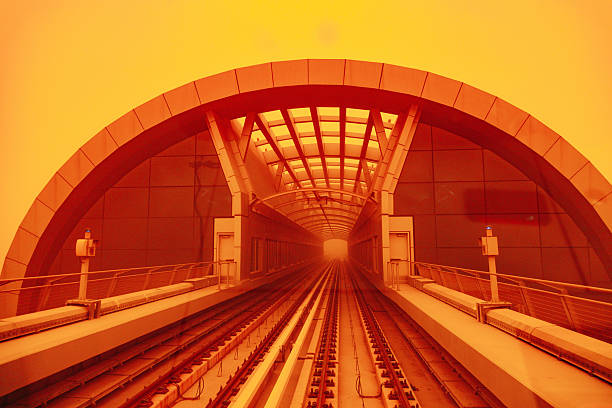 Symmetrical view of railway tracks entering a tunnel bathed in orange light caused by a sandstorm. The heavy dust in the atmosphere creates a surreal and dramatic mood, illustrating how extreme weather can affect transportation systems and visibility in urban environments.