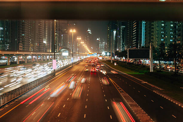 Long-exposure view of nighttime traffic moving along a wide urban highway. Light trails from vehicles illustrate speed, congestion, and the nonstop rhythm of city life, emphasizing transportation demand and urban mobility challenges.