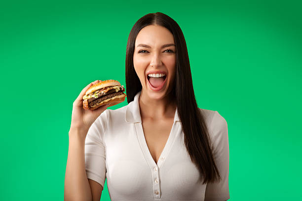 Excited lady eating tasty burger, laughing and looking at camera with open mouth, satisfied woman holding fast food, Isolated over green background