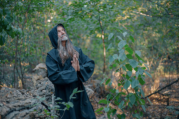 Man with long gray hair and beard wearing a dark hooded cloak praying in a forest