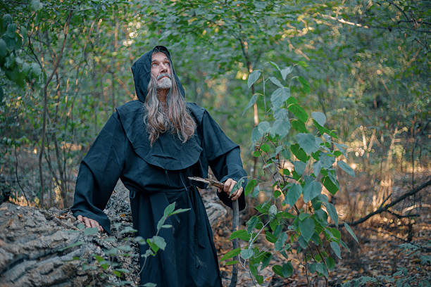 Old man wearing hooded robe standing in a forest while holding a stick