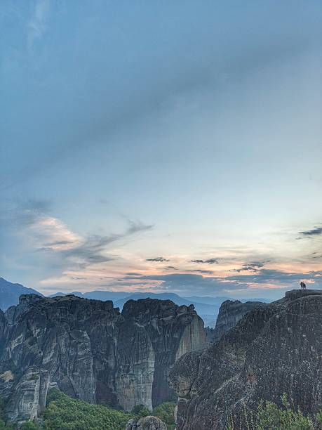 A wide-angle scenic view of the massive sandstone pillars of Meteora in Greece during the golden hour. The image shows the dark, textured rock formations under a soft blue and orange dusk sky, with a small silhouette of people standing on a distant cliff edge. The Pindus Mountains are visible in the misty background.