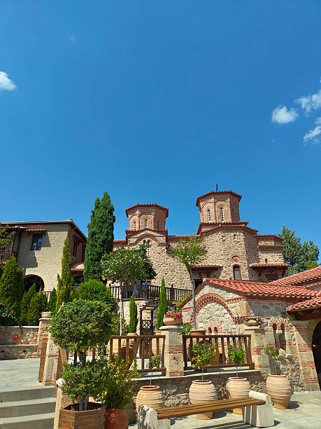 ​A scenic view of a stone watchtower at the Skopje Fortress (Kale) featuring the national flag of North Macedonia. In the background, the modern architecture of the capital city and the slopes of Mount Vodno are visible under a clear blue sky, illustrating the blend of ancient history and contemporary urban life.