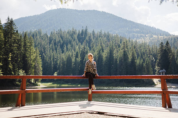 A young woman sits casually on a wooden railing at an observation deck, looking out over mountain lake