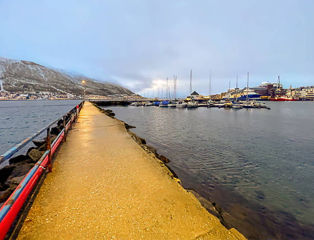 Tromso, Norway - December 23, 2025: Long narrow Loop pier leading to Lokta cabin at end with small yacht docks. Mainland mountain and cityscape