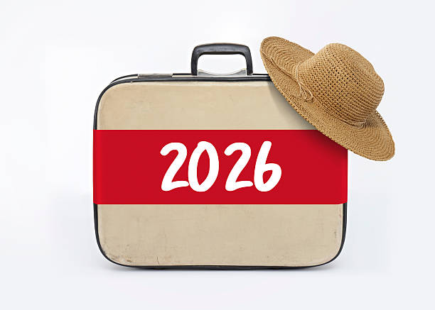 A beige vintage style suitcase with a red 2026 label is topped with a straw hat and placed against a clean white background, symbolizing future vacations, travel goals, and holiday planning. The simple travel still life is ideal for tourism campaigns, trip savings concepts, vacation blogs, and new year resolutions. Horizontal composition with copy space suitable for posters, ads, and editorial layouts.