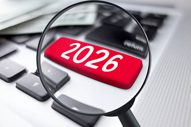 A silver laptop keyboard with a single red 2026 key is viewed through a magnifying glass, emphasizing focus on the coming digital year and technology trends. The conceptual office tech scene is ideal for IT strategy, cyber security, online business planning, and future innovation themes. Horizontal composition with copy space suitable for presentations, reports, and social media posts.