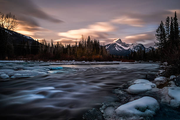 Soft sunrise light illuminates a snow-covered mountain peak in Kananaskis, Alberta, Canada, as a partially frozen river flows through a winter forest landscape under dramatic, streaked clouds. Long exposure utilized in this image to make water flow silky.