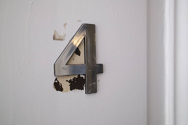Close-up of metallic number four mounted on a white door with peeling paint underneath. Minimalist composition featuring textured surface, weathered detail and residential building entrance concept.