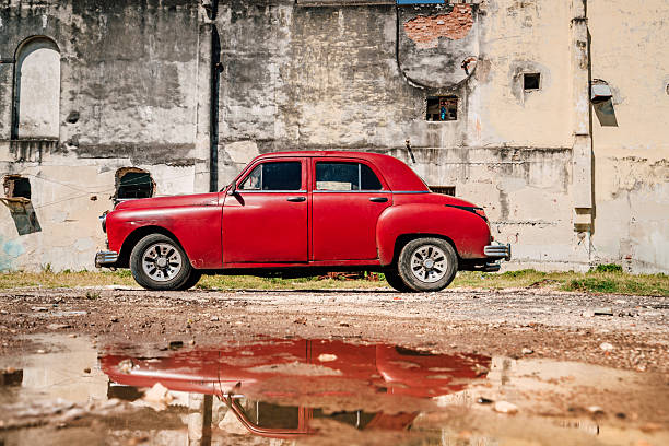 A classic red vintage car sits beside a weathered Havana wall, reflecting in a muddy puddle. The scene blends retro charm with urban decay, capturing Cuban street atmosphere.