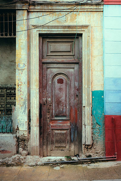 A worn wooden door set in a faded, colorful Havana facade. Cracked plaster, exposed bricks, and hanging wires evoke urban decay and historic charm in Cuba.