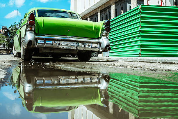 A bright green vintage car sits on a sunny Havana street, its chrome bumper mirrored in a large puddle. A vivid green construction barrier adds bold color to the urban scene.
