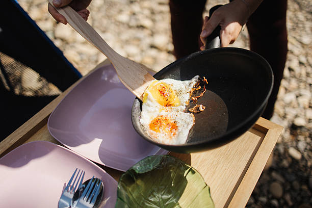 Close-up of an unrecognized person transferring freshly fried eggs from a frying pan onto a plate while cooking outdoors at a campsite.