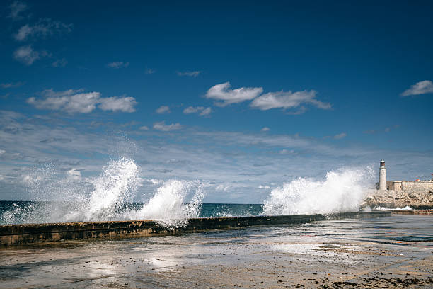 Bold waves crash against a stone seawall near a historic harbor, with a distant lighthouse and blue Havana sky. Dynamic seascape evokes power, travel, and coastal atmosphere.