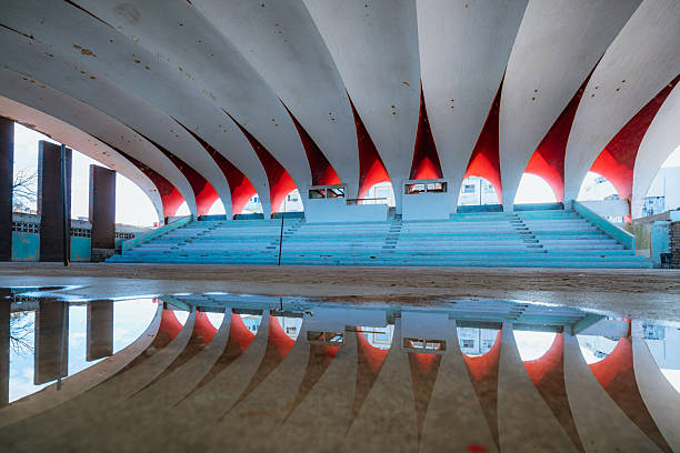 Dramatic curved arches form a stadium-like pavilion with red glow and blue seating, reflected in a tranquil puddle for a symmetrical urban composition. A striking architectural landscape in Havana.