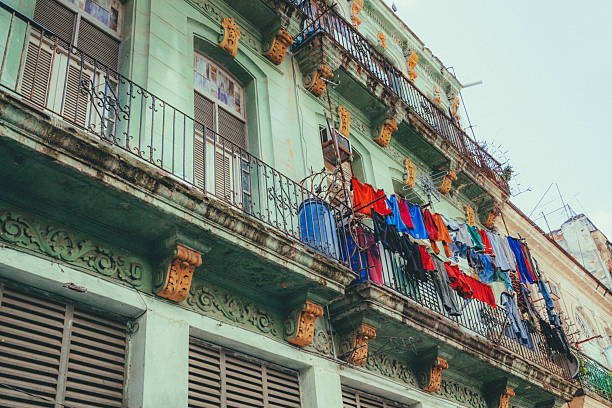 A weathered colonial-style building in Havana, Cuba with ornate balconies, peeling paint, wooden shutters, and colorful laundry hanging on clotheslines in an urban setting