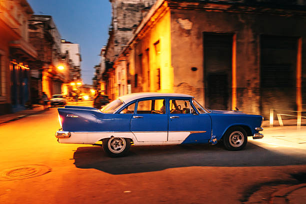 A classic blue and white vintage car cruises a dimly lit Havana street at dusk, exuding retro charm and urban warmth. Warm streetlights and weathered buildings evoke a nostalgic, cinematic city moment.