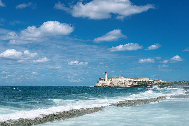 Sunlit fortress and lighthouse rise along the Havana coast, with waves crashing on rocky shore and a clear Caribbean sky. A classic seascape blending history, travel, and coastal beauty.