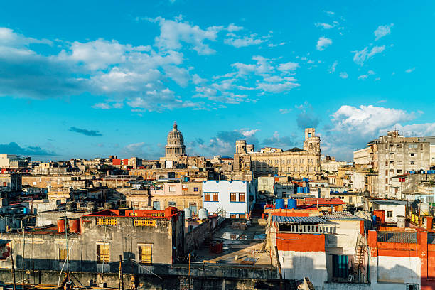 A vibrant Havana cityscape shows worn rooftops and colorful buildings with the National Capitol looming in the distance beneath a bright blue sky. Warm urban atmosphere, historic architecture, and Cuban charm.