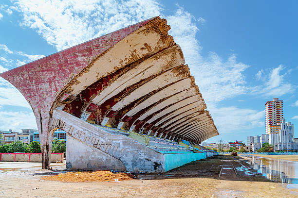A weathered concrete grandstand in José Martí Stadium sits along the field in Havana, Vedado. The open-air bleachers curve toward the city skyline under a bright, blue sky, evoking history and change.