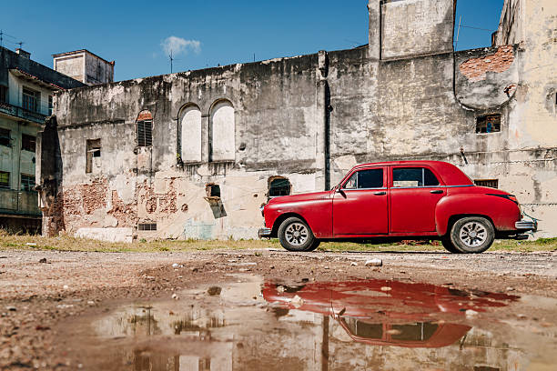 A vintage red car sits beside a weathered concrete wall in Havana, Cuba. The scene blends nostalgia, urban decay, and classic car charm with a reflective puddle in the foreground.