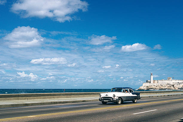 A vintage white-and-black car cruises along a coastal highway in Havana, Cuba, with the sea on one side and a historic fortress and lighthouse in the distance under a bright blue sky.
