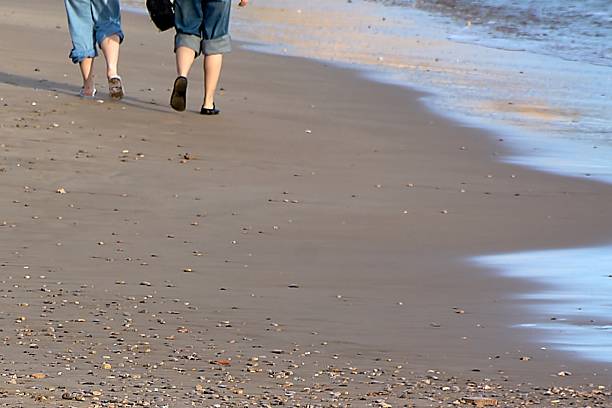 Unrecognizable couple walking along sandy beach shoreline with gentle waves, lifestyle travel concept featuring relaxation, summer vacation, and peaceful coastal escape
