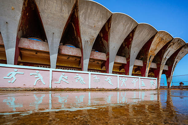 Weathered stadium arches in Havana, with a pink wall and white running figures. Wet ground creates reflections, evoking nostalgia and urban history amid striking architectural shapes.