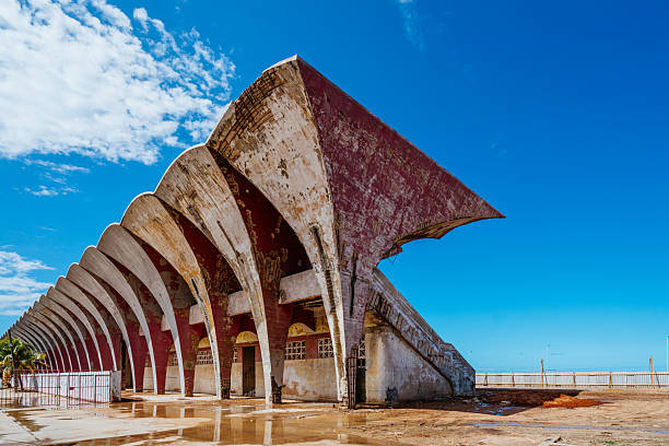 Weathered remnants of Havana's José Martí Stadium line the coast. Streaked concrete arches rise along a salt-washed shoreline under a clear blue sky, telling a story of history, drift, and resilience.