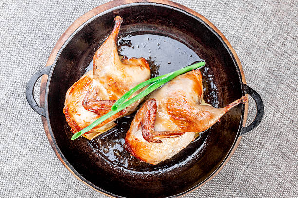 Close-up of two whole fried quails with a crispy golden crust, baked on apple slices. The dish is served hot in a black cast iron skillet on a round wooden base. Garnished with green onions. A rustic style photo focusing on natural ingredients and homemade comfort. View from the top