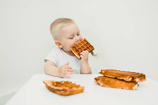 A delightful toddler is seen happily enjoying a delicious waffle treat while sitting at a minimalist white table, showcasing a joyful moment filled with sweetness during breakfast time
