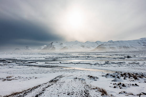 Vast Icelandic winter landscape featuring snow-covered plains and distant mountains under a dramatic sky with sunlight breaking through, showcasing the rugged beauty and isolated nature