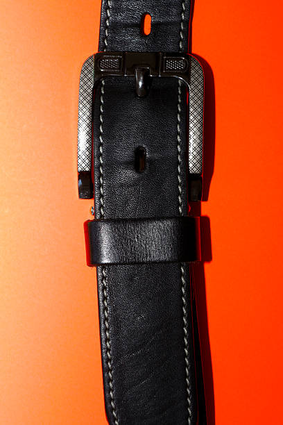 A classic men's black leather belt with a metal buckle decorated with a textured pattern, presented on a bright orange background.