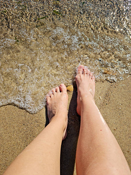 Female feet barefoot in waves of foamy water on a sandy beach on a summer day, top view of female feet, woman on the beach getting her feet wet in warm sea water, carefree vacation by the sea,