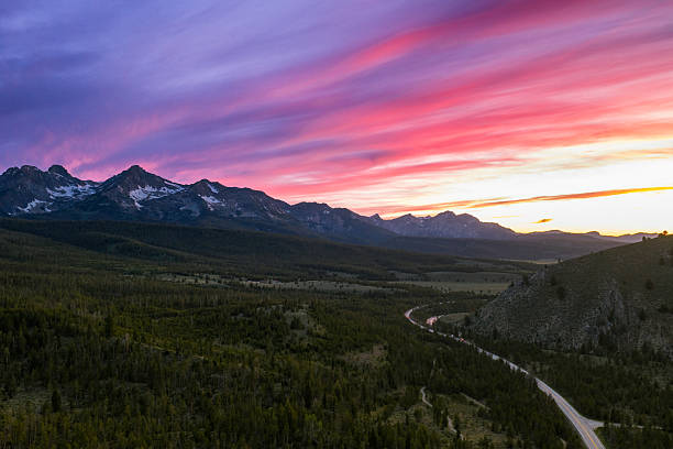 Aerial view of cars traveling along a winding mountain road beside the Salmon River near Redfish Lake in Idaho at sunset as fiery pink and orange clouds stretch across the sky above.