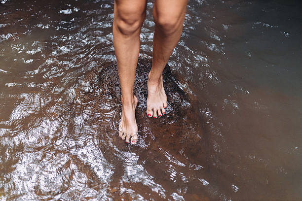 Two bare feet stand at the edge of a shallow, clear river, toes touching wet sand. Calm outdoor scene evokes relaxation, nature, and summertime moments by water.
