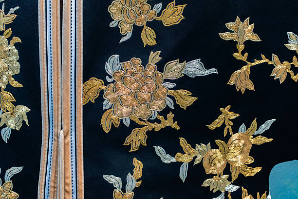 This detailed photograph highlights the intricate embroidery on a piece of dark, luxurious fabric, likely part of a traditional garment. The design features elegant floral motifs—resembling peonies—stitched in shimmering gold and silver threads that create a textured, relief effect against the deep black background. On the left, a vertical hem or opening is visible, trimmed with a light beige border and decorative stitching, emphasizing the fine craftsmanship of the piece.