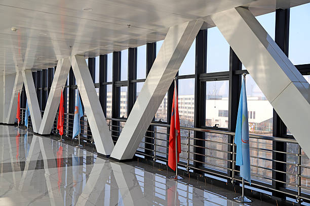 This high-detail, realistic photograph captures a sleek, modern interior corridor featuring dramatic geometric X-shaped metal columns, a highly polished reflective marble floor, and floor-to-ceiling windows that frame urban city views. Vibrant red and blue flags line the walkway, adding pops of color to the contemporary architectural design, while natural light floods the space, emphasizing clean lines and a bright, airy atmosphere. Ideal for corporate, architectural, and interior design-themed projects.