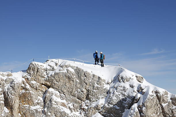 Zugspitze, Germany on November 18, 2025: View of two young mountain climbers who reached the summit of Zugspitze, Germany’s highest peak, on a sunny day. The image captures the alpine scenery, snow-covered peaks, and the achievement of high-altitude mountaineering in the Bavarian Alps.