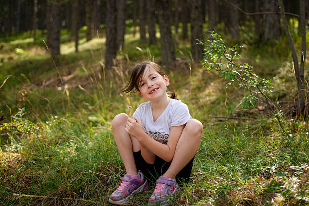 Young girl sitting on green grass in a sunlit forest, smiling at the camera while enjoying outdoor play and carefree summer exploration