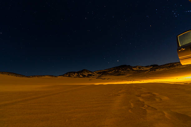 Night desert landscape in Giza, Egypt, with warm artificial light illuminating sand dunes under a starry sky, creating contrast between human presence and vast natural darkness.