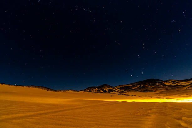 Wide desert landscape in Giza, Egypt, illuminated by warm artificial light beneath a deep starry sky, emphasizing contrast, scale, and the quiet atmosphere of the night desert.