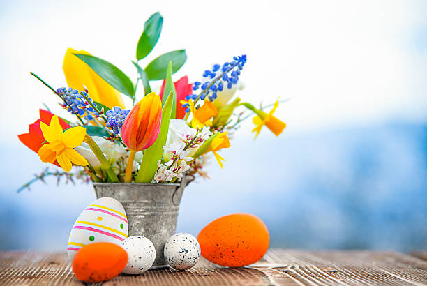 Bright spring still life with a colorful bouquet of tulips, daffodils, and muscari in a rustic metal bucket, decorated Easter eggs on a wooden table, and a soft blue bokeh background with copy space for a seasonal greeting.
