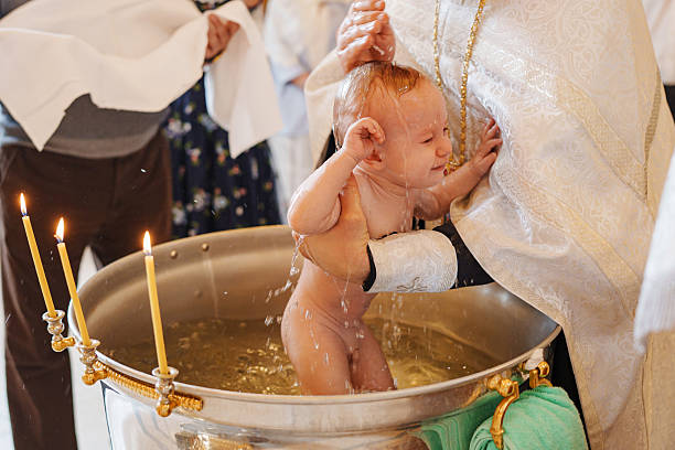A joyful baby is being baptized in a metal basin during a religious ceremony. Family members gather, celebrating this special moment of faith and tradition.