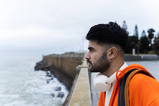 Young adult man wearing orange hoodie and headphones looking at ocean waves
