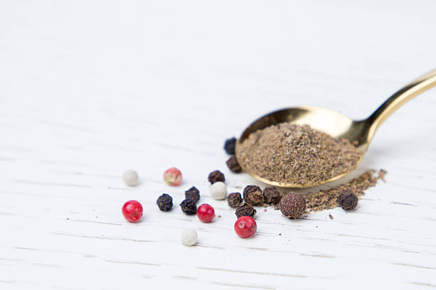 Spoon with pepper powder and peppercorns on white wooden background. Selective focus.