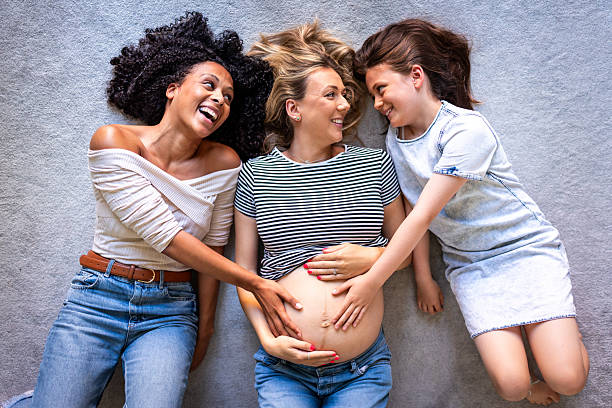 Pregnant woman lying on floor with friends and daughter, all looking at each other, bonding over baby bump