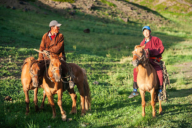 Two riders in traditional clothing sit on chestnut horses in a green, gently sloping valley bathed in warm late-afternoon light. The rider on the left, wearing a brown jacket and cap, holds the reins of two horses and looks to the side; the rider on the right wears a maroon deel and blue cap and faces forward. Rocky hills and patches of shadow form the background, suggesting a quiet pastoral scene of herding life.