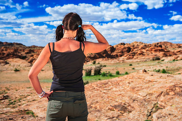 A person seen from behind stands on a sunlit rocky outcrop, shielding their eyes as they look toward a wide expanse of red rock formations and scattered vegetation. They wear a black tank top, olive cargo pants, pigtail braids tied with blue bands, a beaded necklace and bracelets. The scene is bathed in warm light under a vivid blue sky dotted with white clouds, conveying a sense of exploration and open, arid landscape.